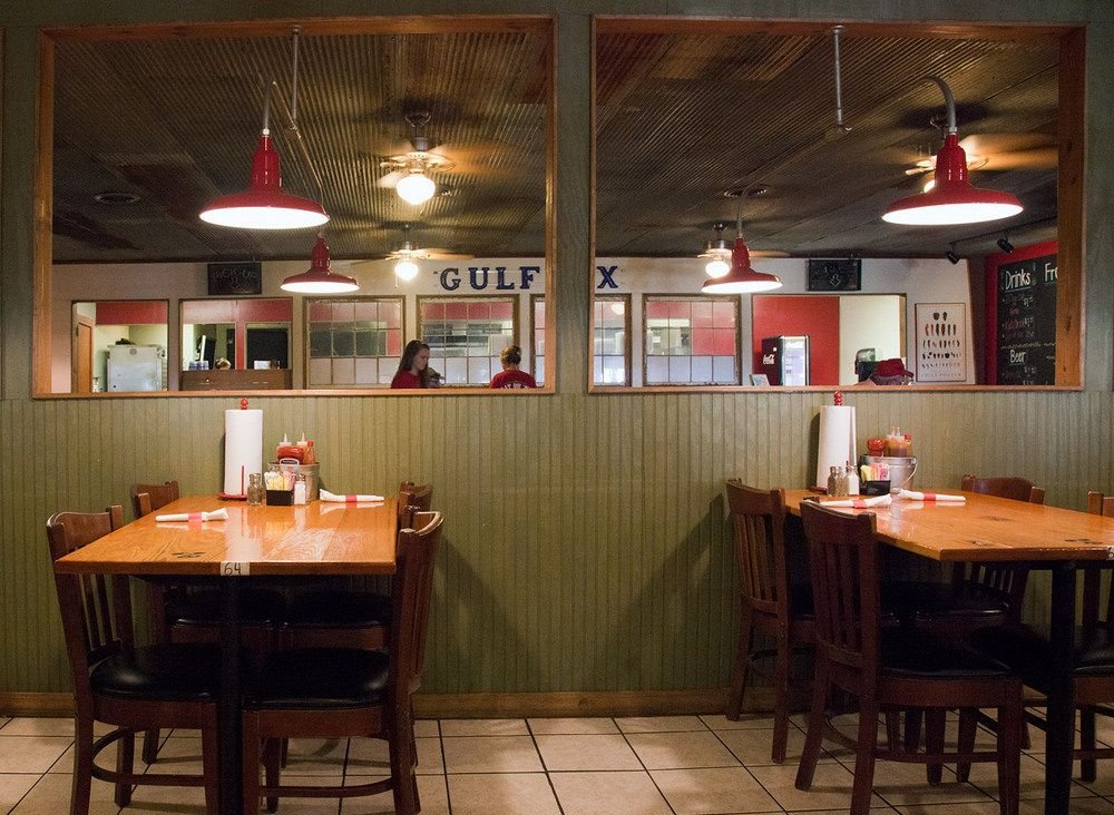 A dining room with wooden tables, red pendant lamps and an old Gulflex sign behind interior windows